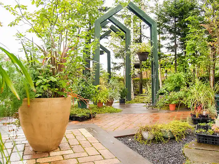 レンガ敷きの通路の先に立つ、家のような形をしたユニークな緑色の木製アーチ。手前には大きなテラコッタの鉢が配置された開放的な空間。