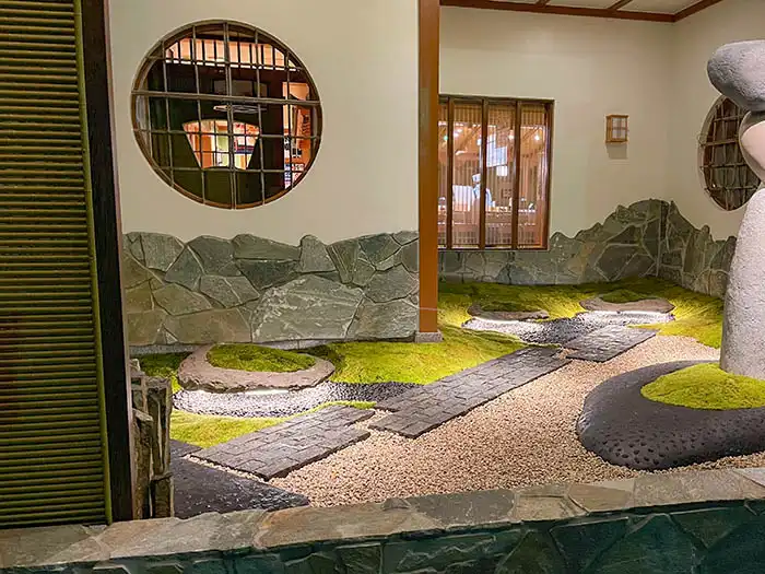 夜の回転寿司店にある半屋内庭園。円窓から光が漏れる白い壁に、石積みのデザインが施され、足元にはライトアップされた苔、砂利、黒い石畳の通路が幻想的に広がる。