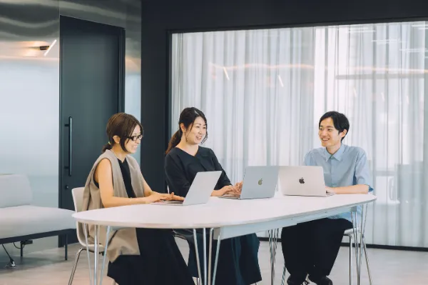 写真。テーブルを囲み、3名の社員が会話をしている様子。手元にはパソコンを開いている