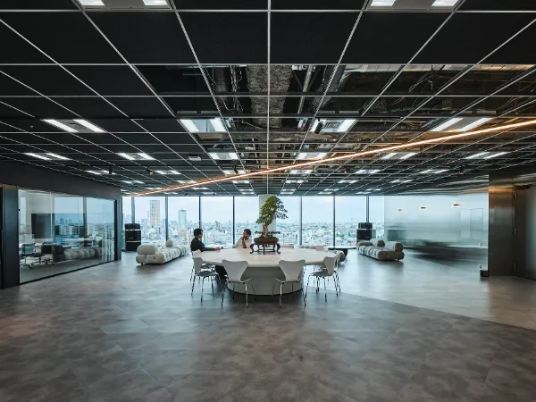 写真、Studioオフィスの様子。中央に円卓が配置され、松の盆栽が設置されている。周囲にはソファスペースや会議室など、自由に利用できる設備が備わっている。天井には、Studioロゴを模した線状の照明が、対角線上に設置されている。