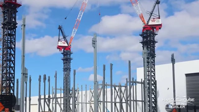 Steel columns being erected at the Gigabay construction site, with two tower cranes actively lifting multiple pieces.