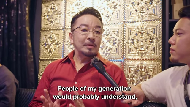 A man in a red shirt (bar owner) being interviewed, gesturing with his hands as he speaks about the district's uniqueness