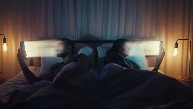 A couple in bed, both looking at their phones, illustrating isolation despite connectivity