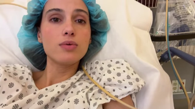 The speaker in a hospital bed, wearing a surgical cap, giving a thumbs up after her egg retrieval surgery