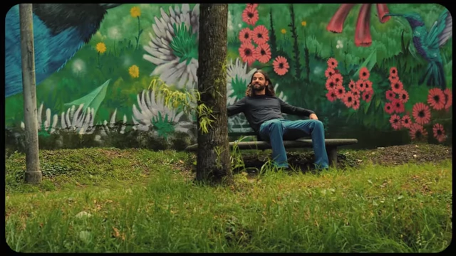 A person sitting on a bench next to a tree with a vibrant mural in the background, depicting a moment of peaceful contemplation in nature, fueling online presence.