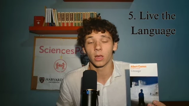 The speaker holds up the French novel "L'Étranger" by Albert Camus, mentioning its usefulness for vocabulary and French exam preparation.
