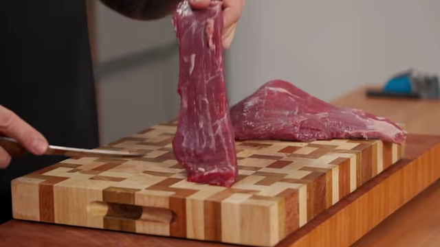 A close-up shot of raw beef showing visible muscle fibers, with the chef demonstrating how to cut across the grain for tenderness