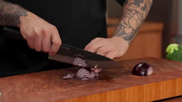 A close-up of a red onion being sliced thinly using the "claw" grip, with the knife resting against the knuckles