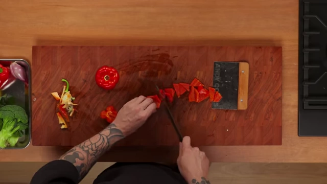An overhead view of a red bell pepper being cut into small, even diamond shapes on a wooden cutting board