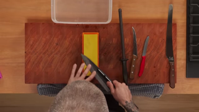 An overhead shot shows the hands of the chef pushing a knife away from himself along a wet stone