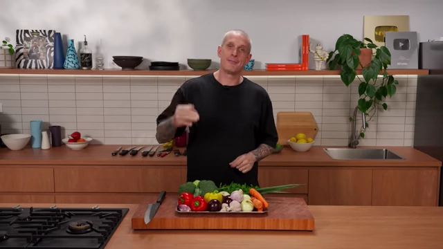 An assortment of fresh vegetables including peppers, onions, garlic, carrots, broccoli, and herbs, displayed on a tray