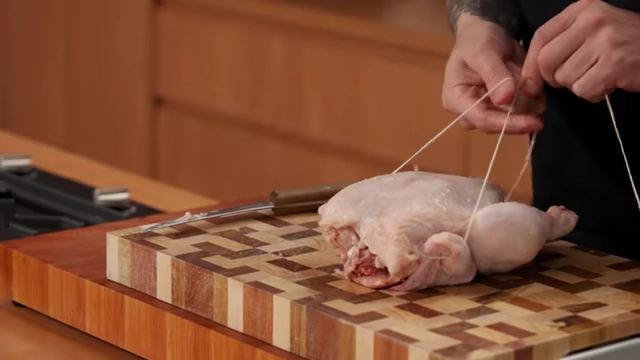 A chef brings butcher's string down around the legs of a raw chicken for trussing