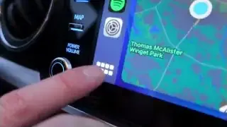 The user's finger points to the grid icon on the Apple CarPlay interface to activate Siri