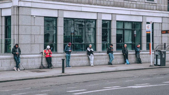 A row of people standing on a sidewalk, many leaning against a wall or other objects, demonstrating widespread poor standing habits.