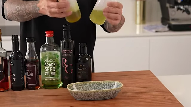 Andy holding squeeze bottles of olive oil and grapeseed oil, demonstrating his preferred method for easy daily use in the kitchen, with various other bottled oils and vinegars arranged behind them.