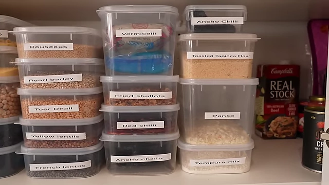 A shelf with labeled containers of various dry ingredients including couscous, lentils, different types of chilies (ancho, red), and baking items like panko and tempura mix.