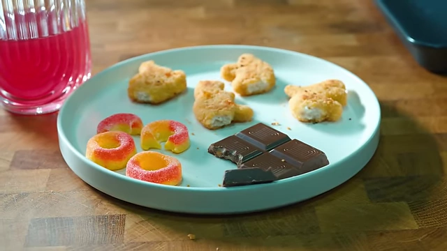 A platter showing MN's limited diet: dinosaur-shaped chicken nuggets, candy gummies, chocolate bar, and a glass of juice.