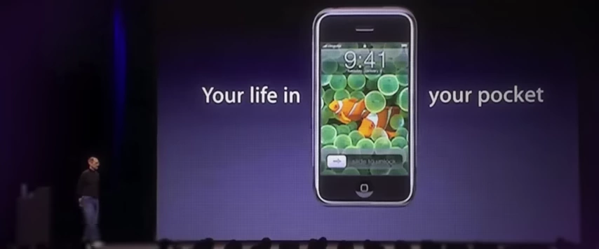 史蒂夫·乔布斯在演示初代iPhone，屏幕上显示“你的生活装在口袋里”。