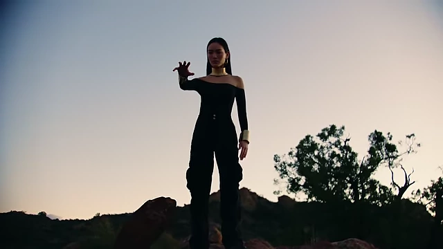 A wide shot shows a woman standing on a hill with multiple dark rocks floating around her against a cloudy sky.