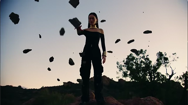 A woman in black stands on a rock formation as dark, jagged rocks float in a circle around her against a cloudy sky.