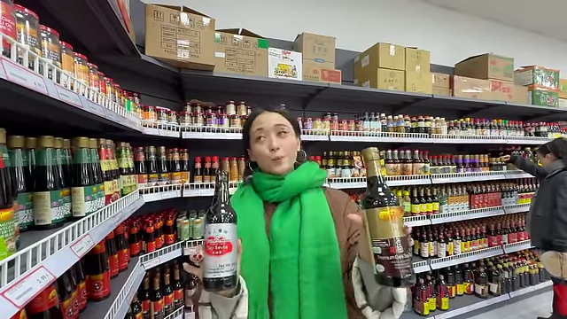 The speaker holding two large bottles of light and dark soy sauce in a grocery store aisle, discussing regional differences.