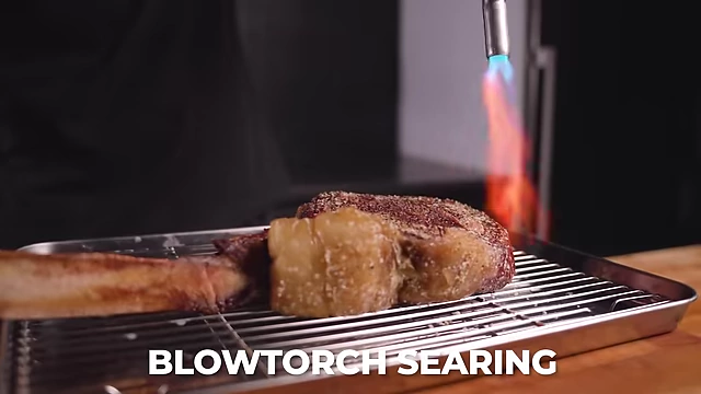 A Tomahawk steak being seared with a blowtorch, demonstrating an aggressive searing technique to develop a crust.