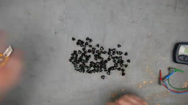 A pile of 98 small black germanium transistors are spread out on a grey surface next to a transistor tester.