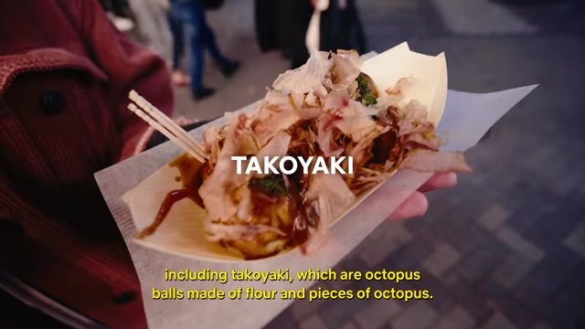 A close-up of freshly prepared takoyaki (octopus balls) garnished with sauce and bonito flakes, served in a small paper boat.
