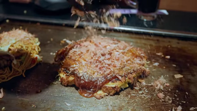 A freshly cooked okonomiyaki, a savory Japanese pancake, topped with sauce, mayonnaise, and green onions on a griddle.