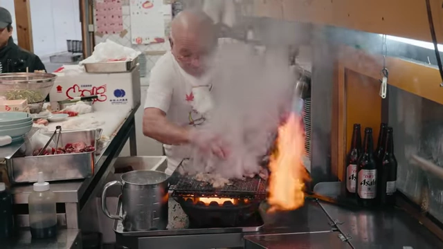 The owner of Izakaya Toyo, Toyo, expertly preparing food on a grill with dramatic flames.