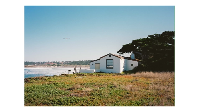 A small white house by the ocean on a grassy hill under a bright blue sky
