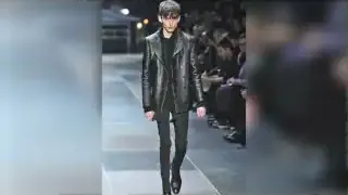 A male model on a runway, showcasing a slim, dark outfit with a leather jacket.