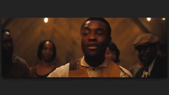 A man speaking in a dimly lit room with others in the background, a scene from "Sinners" suggesting a focus on blues and black culture.