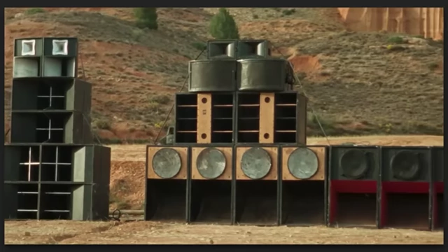 A towering stack of large audio speakers in a desert landscape, central to "Sarat's" sonic experience.