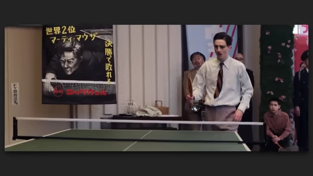 A man in glasses with a mustache, likely Timothy Chalamet, playing ping-pong in a formal setting from "Marty Supreme."
