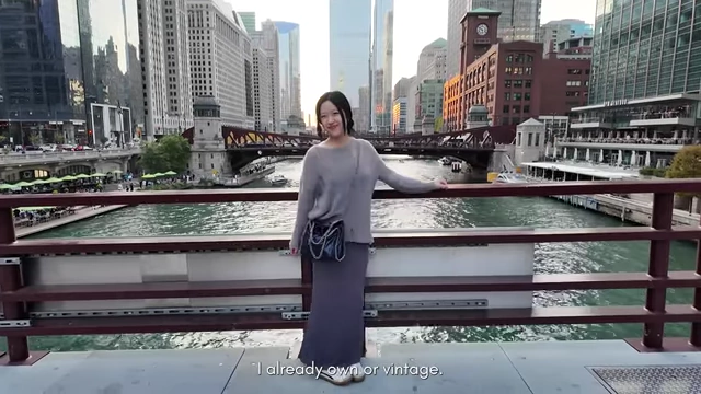 A person in a coat and jeans with a cross-body bag standing on a bridge over a river in a city.