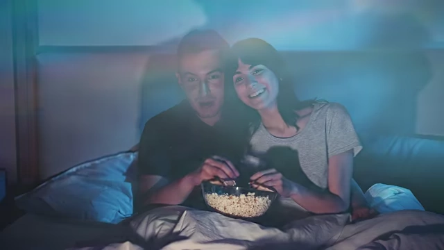 A couple watching TV in bed, illuminated by blue light, which can disrupt melatonin production.