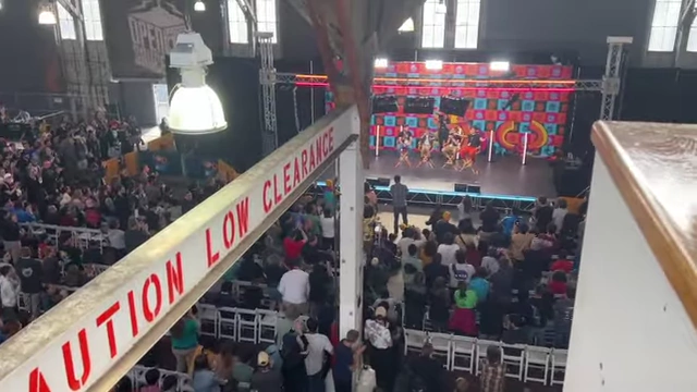 An overhead view of the main stage and crowd in the convention hall, with a "CAUTION LOW CLEARANCE" beam.