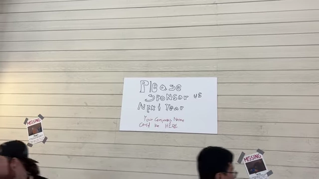 A sign on a wall that reads "Please Sponsor Us Next Year, Your Company Name could be HERE".