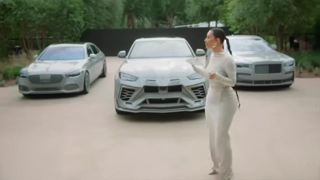 Kim Kardashian standing in front of three luxury cars, all in a matching muted grey color to blend with her minimalist house.