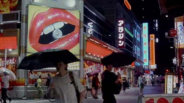 A bustling city street with a large advertisement and people holding umbrellas, indicating crowded conditions due to tourism.
