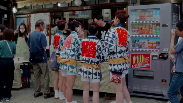 People participating in a local festival, with vendors and vending machines in the background.