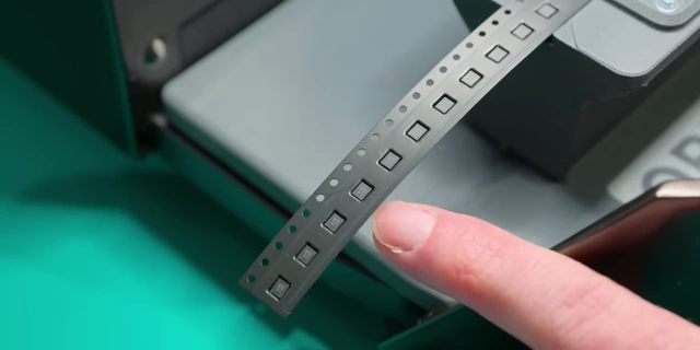 A finger pointing to minuscule SMD components on a tape, highlighting their small size.