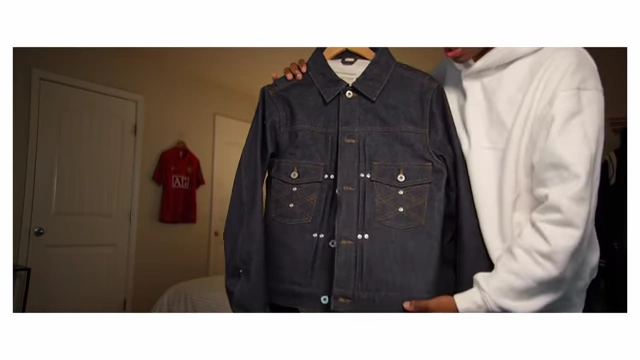 A dark denim jacket with front pleats and two chest pockets, held up by hands