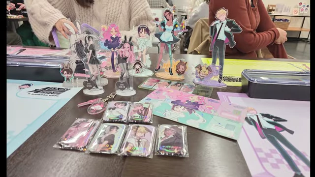 A table covered in various Alien Stage merchandise, including acrylic stands, keychains, and coasters, brought by fans.