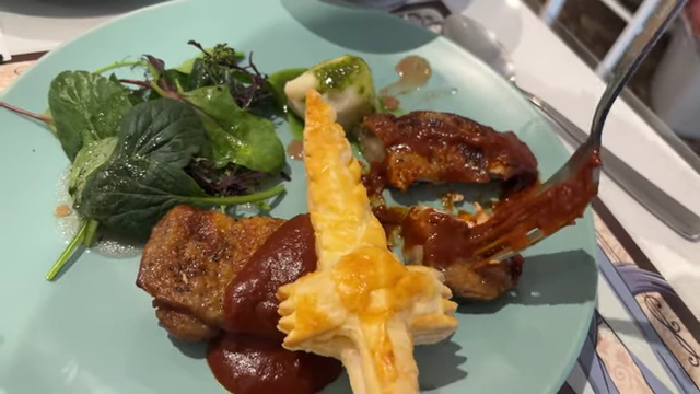 A chicken dish with a sword-shaped puff pastry and a red sauce, served with greens on a light blue plate.