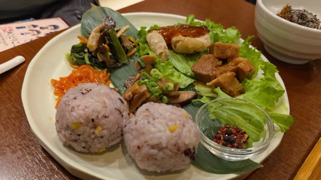 A plate with two rice balls, various vegetables, white fish, and pork belly, served with a small dish of red spices.