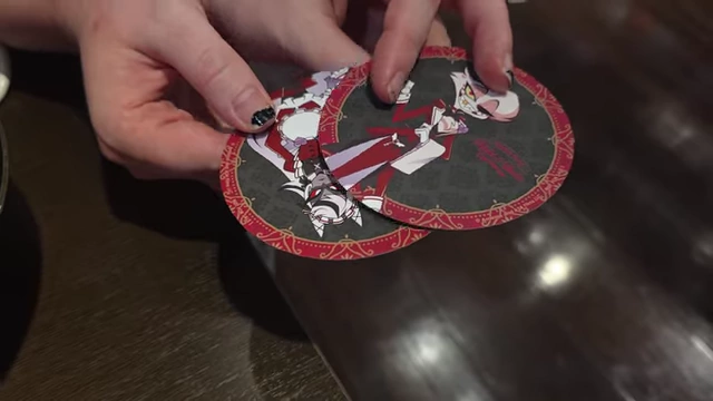 Hands holding multiple round coasters with various Hazbin Hotel character designs.