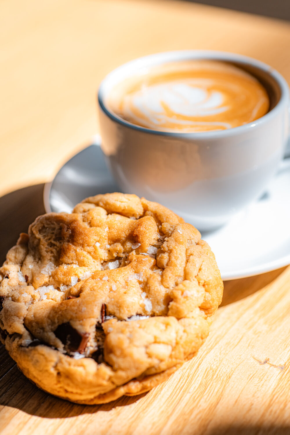 A chocolate chip cookie sitting next to a latte on a blonde table.