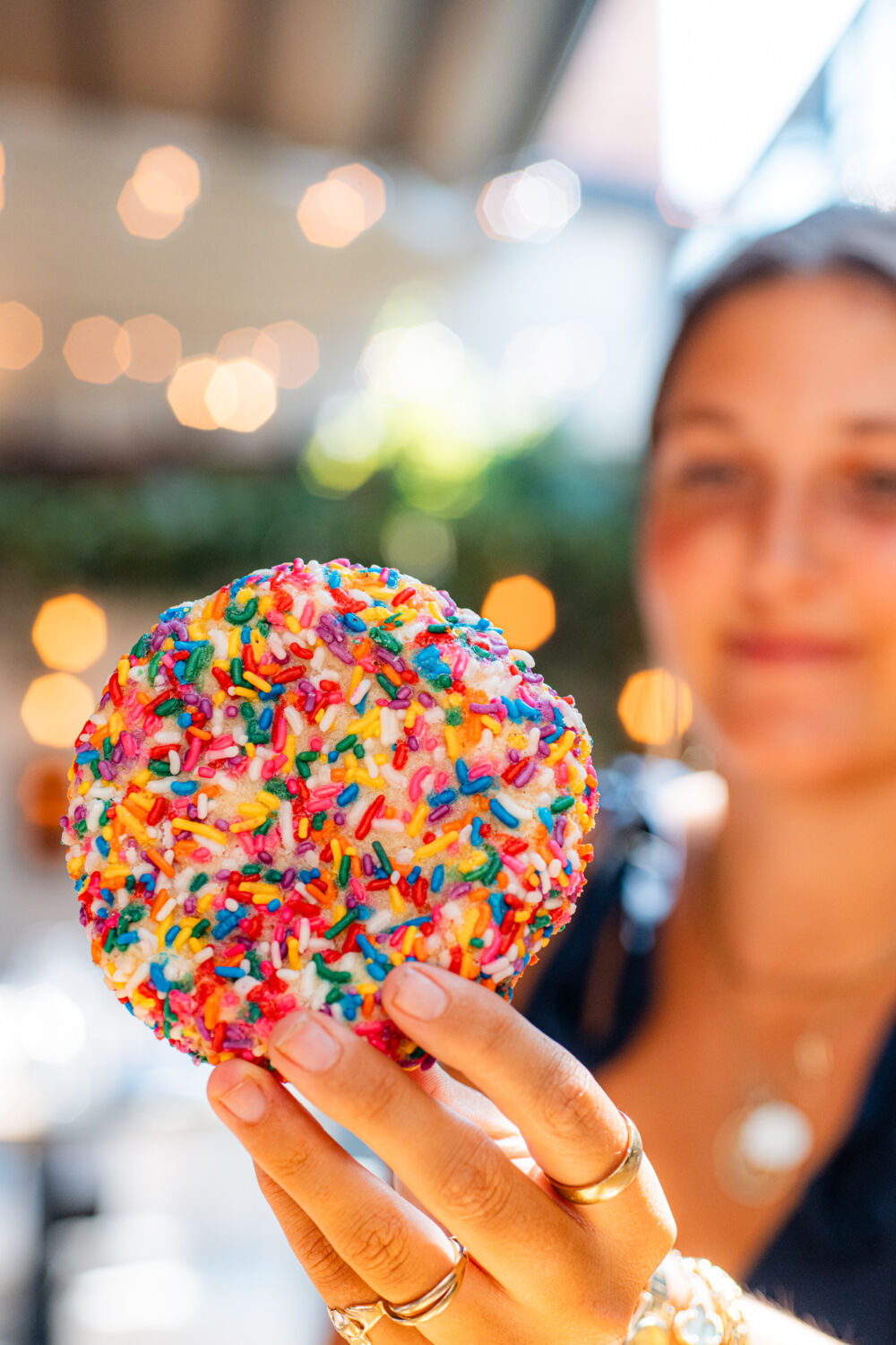 A woman smiling holding a sprinkle cookie with blurred lights and greenery in the background